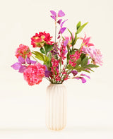 Een crèmekleurige, geribbelde vaas van keramiek bevat het Zijden kunstbloemen boeket Rosy Saga Mist, met roze, rode en paarse zijden bloemen met groene bladeren tegen een lichte achtergrond.