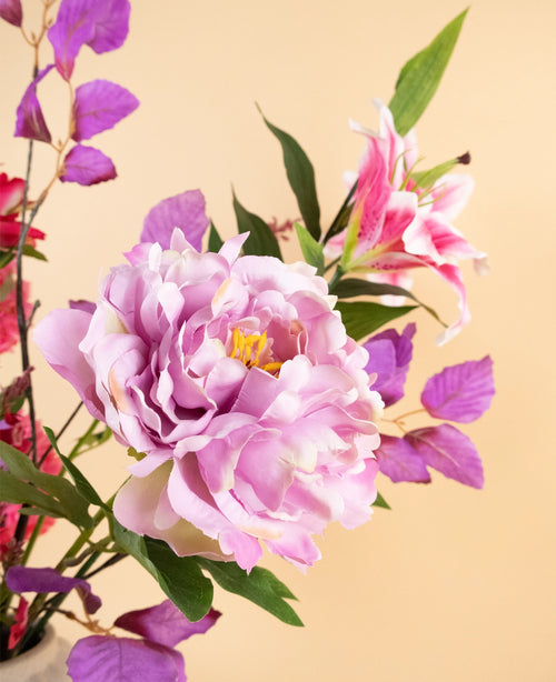 Een close-up van het Zijden kunstbloemen boeket Rosy Saga Mist, met een grote lichtpaarse pioenroos, roze lelie en paarse bladeren tegen een zachtbeige achtergrond.