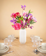 Een witte vaas van keramiek met het Zijden kunstbloemen boeket Rosy Saga Mist fleurt een houten eettafel op voor vier personen met witte borden, kommen, kopjes en glazen, tegen een effen beige muur.