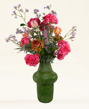 Een groene geribbelde vaas bevat het Zijden kunstbloemen boeket Soft Spring-roze pioenrozen, oranje tulpen, paarse wilde bloemen en groen-duurzaam handgemaakt door Nederlandse bloemisten, tegen een effen lichte achtergrond.