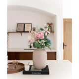 De Vaas Artic Beige Ø21 x H29 cm, gevuld met roze en blauwe kunstbloemen, staat op een zwart boek op een wit aanrecht. Zachte verlichting accentueert het minimalistische tafereel met decorplanken en een houten deur op de achtergrond.