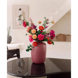 De Vaas Artic Roze Ø21 x H29 cm, gevuld met roze, oranje en rode bloemen, staat op een donker glanzende salontafel in een moderne minimalistische woonkamer met een trap en ingelijste kunstwerken op de achtergrond.
